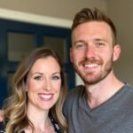 Close-up of a cheerful couple in a casual home environment. The woman has long blonde hair and wears a floral top, while the man has short brown hair and beard, wearing a gray v-neck shirt. Both are smiling warmly at the camera. Behind them is a blue-framed doorway, giving a glimpse of their interior decor.