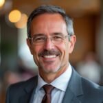 Headshot of a mature businessman with salt-and-pepper hair and neatly trimmed mustache, wearing glasses, a gray suit, light blue shirt, and maroon tie. He's smiling warmly at the camera, exuding confidence and approachability. The background is blurred, showing warm lighting and suggesting a professional office environment.