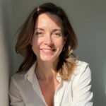 Close-up portrait of a smiling woman with shoulder-length brown hair, wearing a crisp white shirt. She's leaning against a light-colored wall, with sunlight casting shadows, creating a warm and inviting atmosphere. Her genuine smile and relaxed posture convey confidence and approachability in a professional setting.