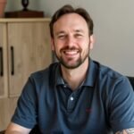 Smiling man with short brown hair and beard wearing a dark blue polo shirt, seated in a home office setting. Behind him are light wooden cabinets. He exudes warmth and approachability with his genuine smile and relaxed posture, suggesting a professional yet comfortable work environment.